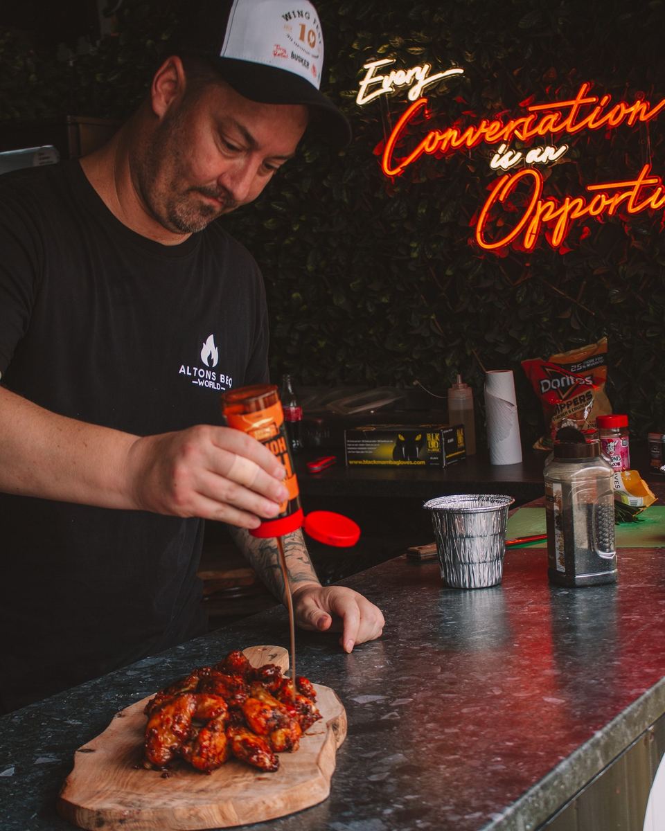 Chef drizzling hot honey over wings beneath a neon sign